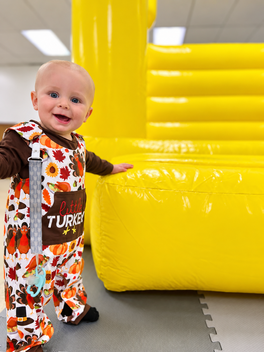 Smiling toddler in a Little Turkey fall outfit standing beside a big yellow inflatable at an indoor celebration