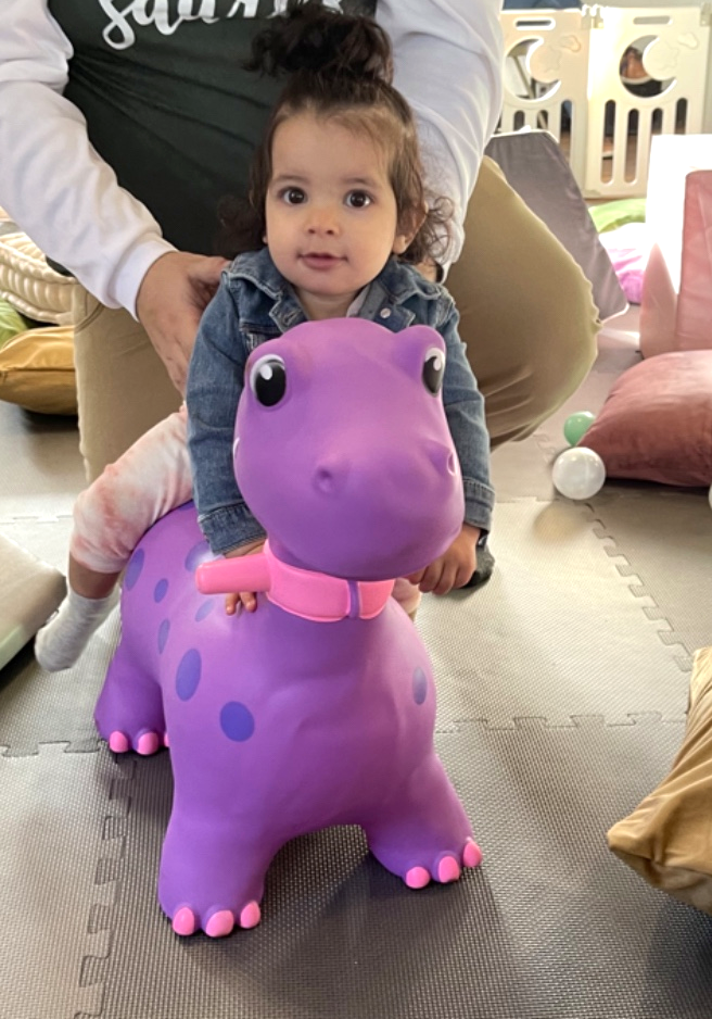 A toddler girl riding a purple hopper toy on the soft play mats