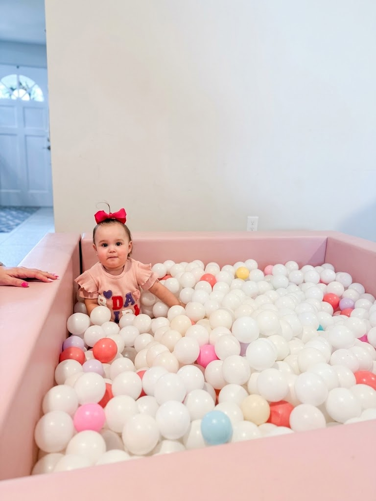 Baby girl with a red bow smiling in a pink square ball pit filled with white and pastel balls