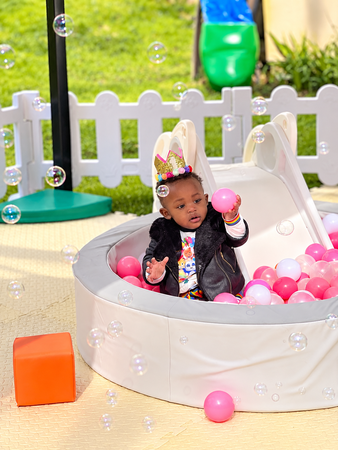 Birthday girl in a crown playing with a pink balloon inside the white round ball pit with bubbles outdoors